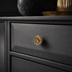 Puede incluir: Un pomo de armario circular con un borde dorado metálico y una incrustación central con un patrón de mármol veteado en tonos marrón y negro. El pomo está montado en un frente de cajón de madera negro mate con veta visible y bordes biselados.