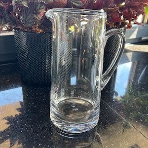 May include: A clear glass pitcher with a handle and a pouring spout. The pitcher is empty and reflects the surrounding environment. The pitcher is sitting on a dark surface, with potted plants in the background.