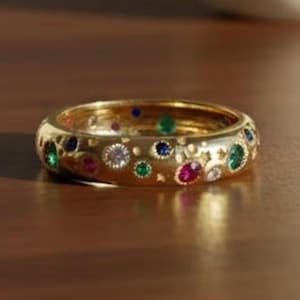 Peut inclure: Une bague en or ornée d'une bande ornée de diverses pierres précieuses colorées, notamment des pierres vertes, bleues, rouges et claires. La bague présente un design détaillé avec de petites découpes, sur un fond brun chaud.