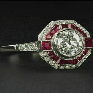 Peut inclure: Bague de style Art Déco avec un grand diamant rond et clair en son centre. La bague est entourée de pierres précieuses rouges et de petits diamants, sertis dans un métal argenté. La bande est également ornée de petits diamants.