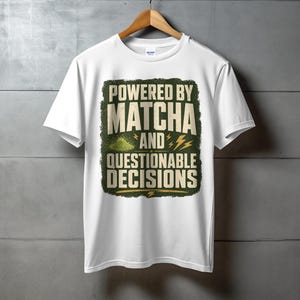 May include: White t-shirt with the text "POWERED BY MATCHA AND QUESTIONABLE DECISIONS" in a green and yellow design. The shirt is hanging on a wooden hanger against a grey wall.