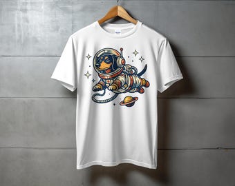 May include: White t-shirt featuring a cartoon dachshund in an astronaut suit, floating in space with a planet. The dog is wearing a space helmet and suit with orange, brown, and blue accents. The design includes stars and a small planet.