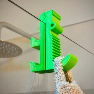 Puede incluir: Un gancho verde brillante para puerta de ducha, diseñado para sujetar una toalla. El gancho es de plástico y tiene un diseño único con un agarre texturizado y dos brazos. La toalla es de color marrón claro.