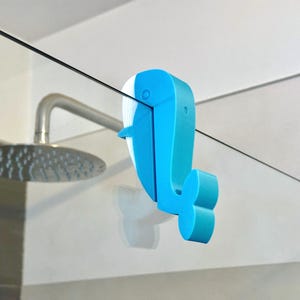 Könnte beinhalten: Ein blau-weißer Wal-Duschhaken. Der Haken besteht aus Kunststoff und ist so konzipiert, dass er über den Rand einer Glas-Duschabtür gehängt werden kann. Im Hintergrund ist ein Duschkopf zu sehen.