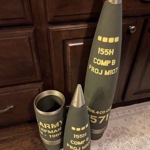May include: Three olive drab artillery shell components with yellow stenciled text. The tallest piece is a complete projectile marked 155H COMP B PROJ M107. A smaller projectile and a cylindrical base section are marked ARMY LEFMAN 1966.