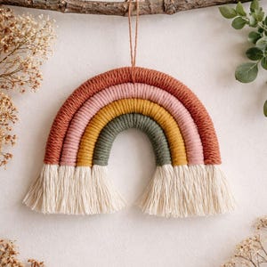 Könnte beinhalten: Ein handgefertigter Makramee-Regenbogen zum Aufhängen. Der Regenbogen besteht aus Garn in Terrakotta, Rosa, Gelb und Grün, mit cremefarbenen Fransen. Er hängt an einem natürlichen Ast.