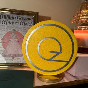 May include: A yellow and silver decorative lamp with a stylized "Q" design. The lamp sits on a gold base. A framed vintage newspaper print and a lit candle are in the background.