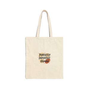 May include: Cream-colored tote bag with a simple design. The bag features a pair of handles and the words "PERFECTLY IMPERFECT HUMAN" in a playful font with orange and yellow accents. The bag is made of a sturdy fabric.