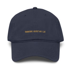 Può includere: Cappellino da baseball blu navy con visiera curva. Il cappellino presenta la scritta ricamata "THINKING ABOUT MY CAT" in oro sul davanti. Ha una corona arrotondata e un bottone sulla parte superiore.