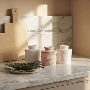 May include: Three ceramic butter crocks with lids, two in solid cream and one with a pink and white marbled finish. Each container features the word Butter embossed on the front. A small plate with sprigs of dried herbs sits in the foreground.