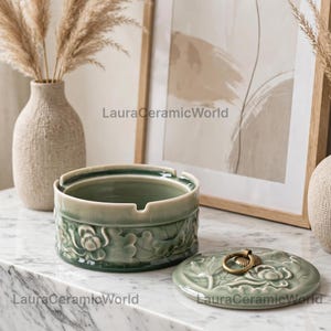 May include: A round sage green ceramic ashtray with a removable lid featuring a brass ring handle. The exterior of the ashtray and the lid are decorated with a raised relief floral pattern. The text LauraCeramicWorld is visible on the image.