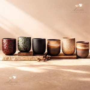 Puede incluir: Colección de seis tazas de cerámica en varios colores y texturas. Las tazas, de forma cilíndrica y base redondeada, se exhiben sobre una superficie de madera. Los diseños incluyen rojo texturizado, beige moteado y marrón a rayas.