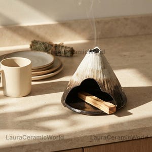 Puede incluir: Un quemador de incienso cónico de cerámica con la parte superior blanca y la base marrón oscuro, con un exterior texturizado y una amplia abertura arqueada. Dentro hay un palo de madera claro y un hilo de humo que sube desde la cima.
