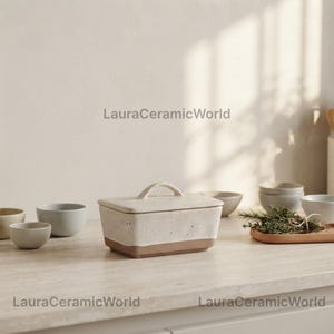 Può includere: Un burriera rettangolare in ceramica con coperchio e manico, caratterizzata da una parte superiore bianco sporco maculata e una base marrone tinta unita. Il testo LauraCeramicWorld è visibile. Diverse piccole ciotole in ceramica e un vassoio in legno con erbe.