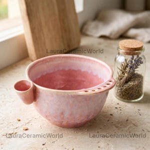 May include: A round ceramic bowl with a glossy pink and white glaze. The bowl features a small conical side attachment and a flat handle extension with five circular holes. The text LauraCeramicWorld is visible in the background.