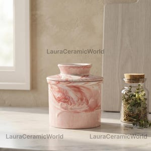 Puede incluir: Un recipiente de cerámica cilíndrico para mantequilla con un patrón jaspeado en rosa y blanco y un borde superior acampanado. El texto LauraCeramicWorld es visible en la imagen. Un frasco de vidrio transparente con hierbas verdes secas a la derecha.