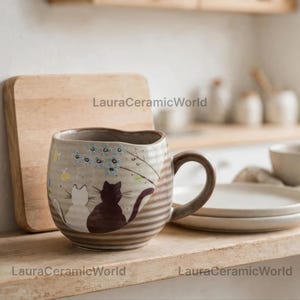Może przedstawiać: Ceramiczny kubek o żebrowanej fakturze z ręcznie malowaną ilustracją przedstawiającą białego i ciemnobrązowego kota siedzących pod gałęzią z jasnoniebieskimi kwiatami i żółtymi akcentami. Kubek posiada zakrzywione ucho i jasnobeżowy kolor podstawowy.