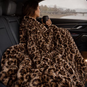 May include: A plush, faux fur throw blanket featuring a brown and black leopard print pattern. The blanket has a thick, textured pile and is draped over the lap, covering the lower body while providing a soft, dense, and fuzzy appearance.