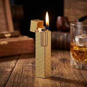 May include: A gold-coloured lighter with a textured, pyramid-like pattern. The lighter is open, with a flame burning. A glass of amber liquid with ice cubes sits nearby on a wooden surface. A cigar box and pipe are in the background.