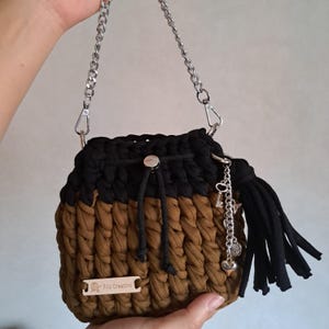 Op de afbeelding: Een gehaakte emmer tas in zwart en bruin met een zilveren kettingriem en een zwarte kwast. De tas heeft een trekkoordsluiting en decoratieve bedels. Een klein houten label leest "Filo Creativo".