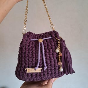 Op de afbeelding: Een handgemaakte, dieppaarse gehaakte tas met een gouden kettingriem. De tas heeft een trekkoordsluiting met een gouden accent, een bijpassende kwast en decoratieve bedels. De tas heeft een klein houten label met de tekst "Filo Creativo".