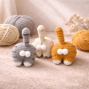 May include: Three handmade crochet cat butt ornaments in gray, cream, and orange. Each has a tail and two white pom-poms. Yarn balls in gray and cream are in the background, suggesting a craft theme.