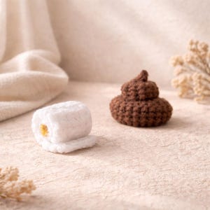 Op de afbeelding: Een witte gehaakte toiletpapierrol en een bruine gehaakte poep-emoji liggen op een getextureerd oppervlak. De toiletpapierrol heeft een gele kern. De poep-emoji is donkerbruin van kleur.