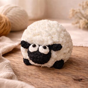 May include: A handcrafted sheep toy, featuring a fluffy white body and black face, ears, and feet. The sheep has large, round eyes. It is placed on a wooden surface. The toy is likely crocheted or knitted.