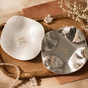 May include: Two irregular, organic-shaped trinket dishes. One is matte white with the text 'b & f Engaged 08.17.2026' in gold. The other is a reflective, metallic silver finish with the text 'a & t 08.17.2026' in gold. Both feature smooth, wavy edges.