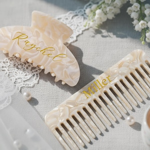 以下が含まれることがあります： 金色の文字が入った明るい色のヘアクリップと櫛のセット。ヘアクリップには「Raychel」、櫛には「Miller」と書かれています。どちらのアイテムもマーブル模様で、グレーの表面にレースと真珠が置かれています。