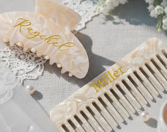 Könnte beinhalten: Eine cremefarbene, marmorierte Haarspange mit dem Namen Raychel in goldener Schreibschrift und ein passender Haarkamm mit dem Namen Miller in goldener Schreibschrift. Beide Artikel haben ein glänzendes, perlmuttartiges Finish.