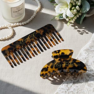 May include: A tortoiseshell-patterned comb and hair claw clip, both personalized with the name "Elizabeth" in gold script. The comb is rectangular with evenly spaced teeth. The clip is a curved shape with a secure clasp.