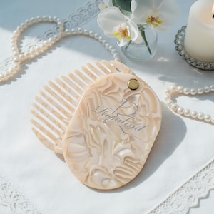May include: A light beige, oval-shaped hair comb with a matching cover. The cover is engraved with the name "Rosalind" in a script font. The comb and cover have a marbled effect. A gold-coloured rivet secures the cover.