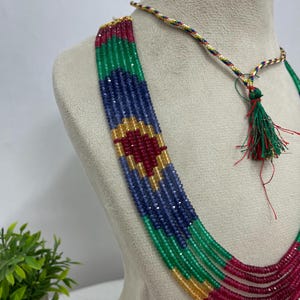 Op de afbeelding: Een meerstrengs ketting met gefacetteerde kralen in levendige tinten rood, groen, blauw en geel. De kralen zijn gerangschikt in een geometrisch patroon en de ketting bevat een decoratief koord met een kleine groene en rode kwastsluiting.