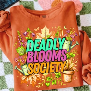 Könnte beinhalten: Ein rostorangefarbenes Sweatshirt mit Rundhalsausschnitt und einem Grafikdruck mit dem Text DEADLY BLOOMS SOCIETY in Türkis, Pink und Orange. Das Design zeigt Illustrationen von Ahornblättern, violetten Glockenblumen und zwei Gießkannen.