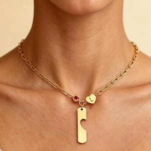 May include: Gold-colored necklace with a rectangular pendant and a heart-shaped charm. The necklace has a chain link design and a small red gemstone. The heart charm has the letter "L" on it.