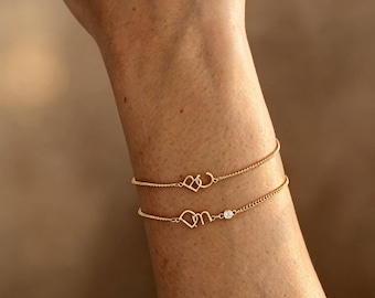 Op de afbeelding: Twee delicate gouden kettingarmbanden. De bovenste armband heeft een kleine hartvormige bedel en een ronde bedel. De onderste armband toont een kleine letterbedel en een enkele ronde heldere steen in een gouden zetting op de dunne ketting.