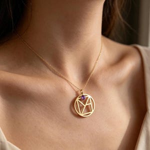 May include: A gold-toned necklace featuring a circular pendant with an openwork geometric design and a single small, round, purple gemstone set at the top. The pendant hangs from a delicate, thin gold-toned chain resting against the collarbone.