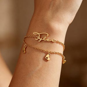 Peut inclure: Un bracelet à double chaîne doré avec une plaque nominative en écriture cursive lisible Tylee. La chaîne inférieure est ornée de trois petits pendentifs en forme de cœur, chacun avec un centre évidé, créant un aspect superposé délicat.
