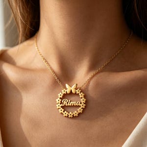 Pode incluir: Um colar de tom dourado com um pingente circular que apresenta uma borda floral e uma pequena borboleta no topo. O nome Rima está escrito em letra cursiva no centro do círculo, suspenso por uma delicada corrente de tom dourado.