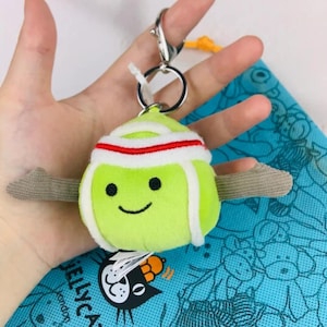 May include: A green plush tennis ball keychain with a smiling face, white trim, and a red and white headband. The keychain has gray fabric arms and a silver clasp. A blue bag with a cartoon design is in the background.