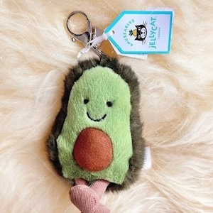 Może przedstawiać: Pluszowy brelok do kluczy w kształcie awokado z uśmiechniętą buzią i brązową pestką. Awokado jest zielone z brązowym, futrzanym obramowaniem. Ma różowe nóżki i srebrny karabińczyk. Etykieta z napisem "Jellycat London".