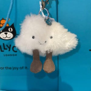 May include: A white plush cloud-shaped keychain with a smiling face and two brown dangling legs. The keychain has a silver clasp and a small tag. The background is a bright blue.