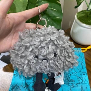 Puede incluir: Un llavero de felpa gris con forma de animal esponjoso de textura rizada. El llavero tiene ojos negros y una boca pequeña cosida. Cuelga de un anillo de metal plateado.