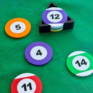 May include: A selection of colourful, circular coasters resembling snooker balls, each with a white number. The coasters are orange, purple, green, and red. A black triangular holder contains more coasters.