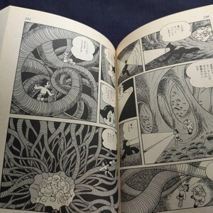 May include: Open book with black and white manga illustrations. Panels depict a science fiction scene with characters in a cavernous environment with large, tentacle-like structures. Speech bubbles contain Japanese text.