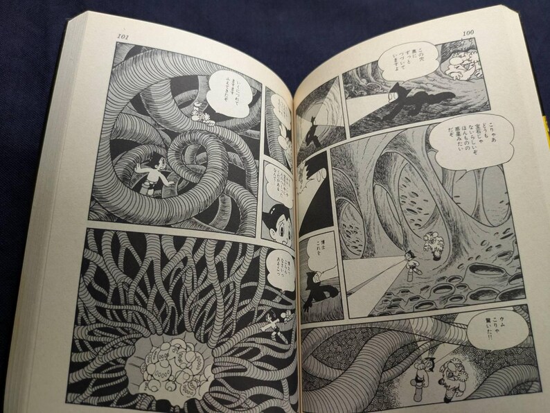 May include: Open book with black and white manga illustrations. Panels depict a science fiction scene with characters in a cavernous environment with large, tentacle-like structures. Speech bubbles contain Japanese text.