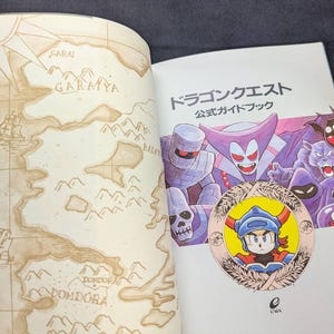 May include: Open book with a map on the left page and a colorful illustration on the right. The right page features the text "Dragon Quest" in Japanese, along with character illustrations.