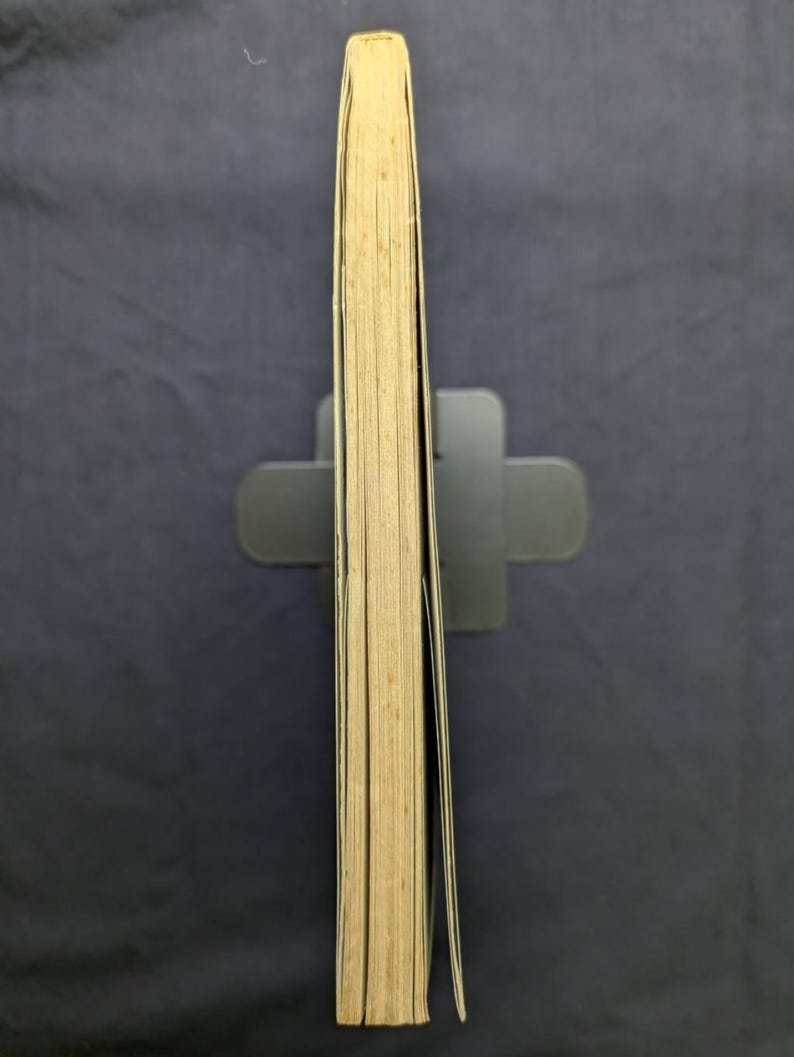 May include: A book with yellowed pages, held upright by a gray book stand. The book's spine is visible, showing the thickness and the aged appearance of the paper. The edges of the pages are slightly yellowed, indicating age.