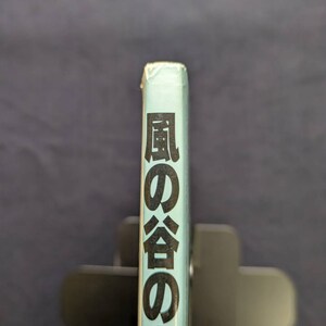 May include: A light blue book spine with Japanese text in black. The book is held in a black book holder against a dark blue background. The text reads "風の谷のナウシカ".